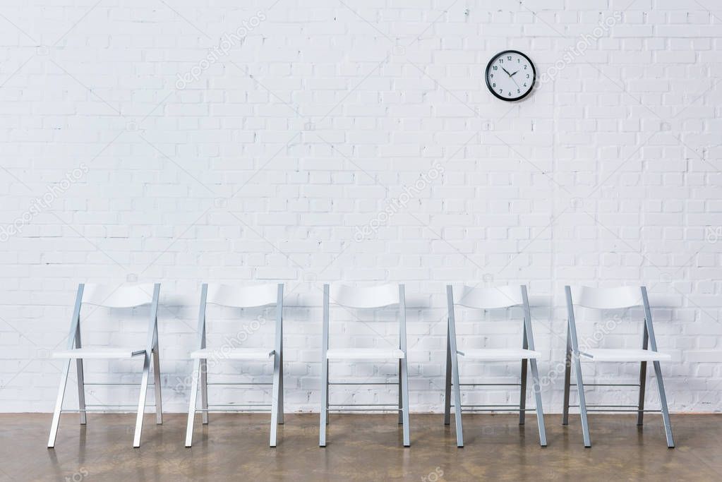 Row of empty chairs by white brick wall with clock