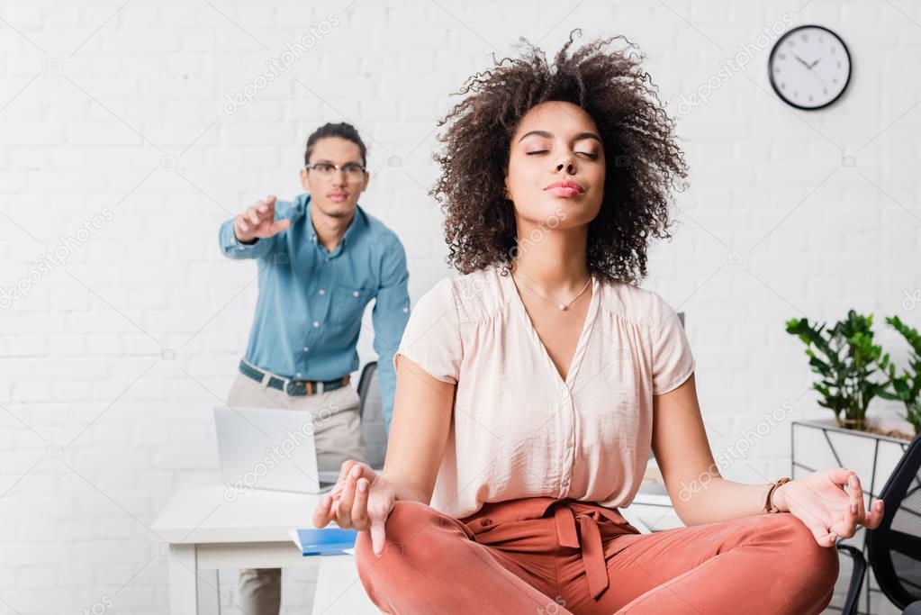 Young businesswoman relaxing and meditating in office with male coworker behind