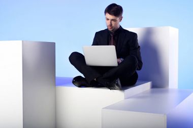 businessman using laptop while sitting on white block isolated on white clipart