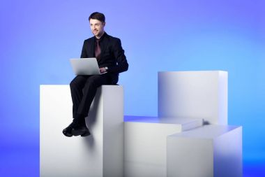 beyaz izole beyaz blokta oturan dizüstü bilgisayar ile gülümseyen iş adamı