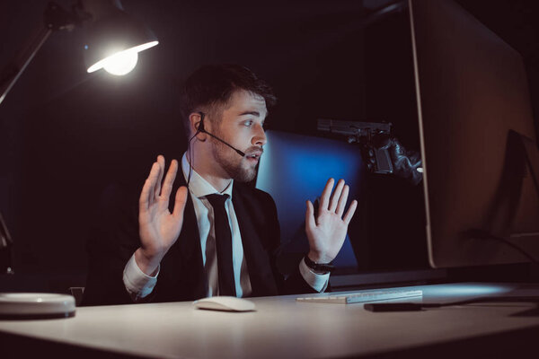 Agent with gun at head and hands up sitting at table with computer screen in dark
