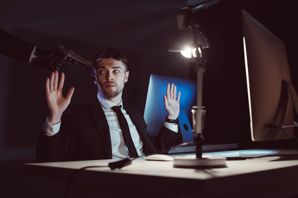 Agent with gun at head and hands up sitting at table with computer screen in dark
