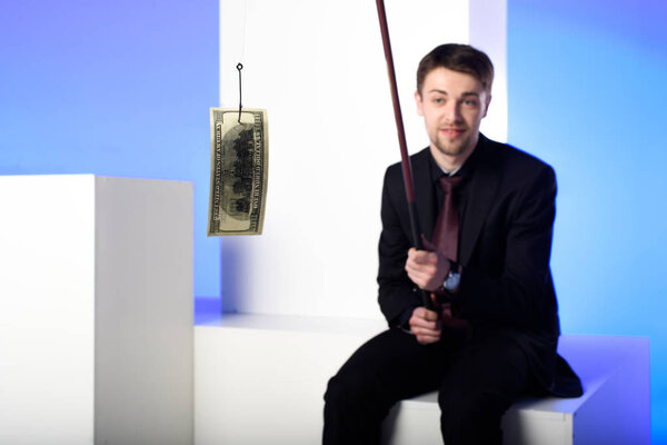 businessman in suit holding fishing tackle with dollar while sitting on white block isolated on blue