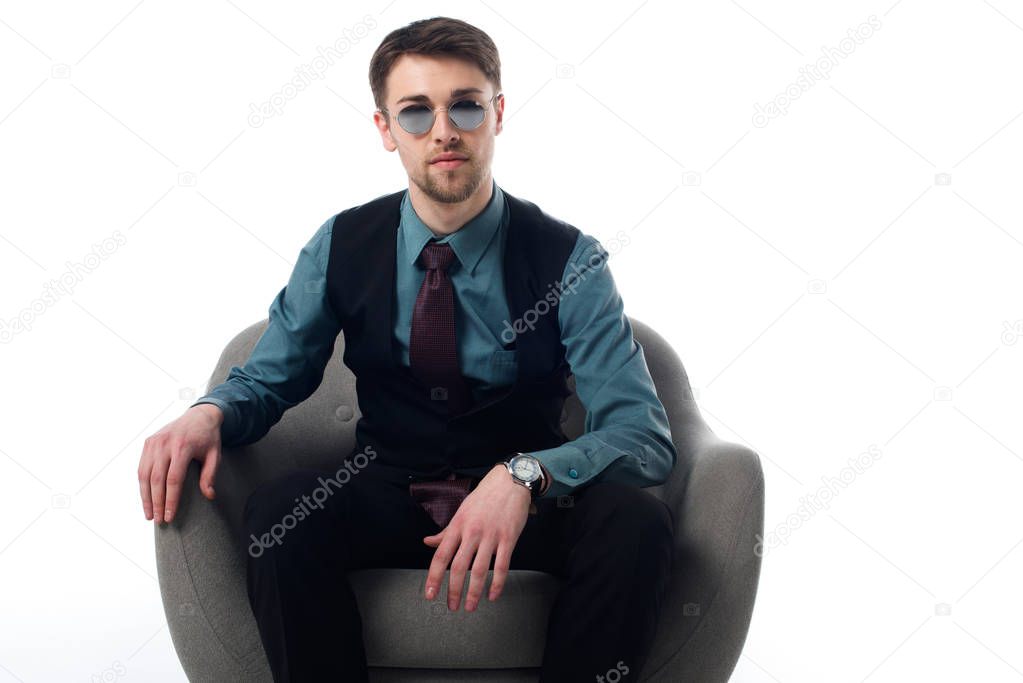 portrait of stylish spy in sunglasses sitting in armchair isolated on white
