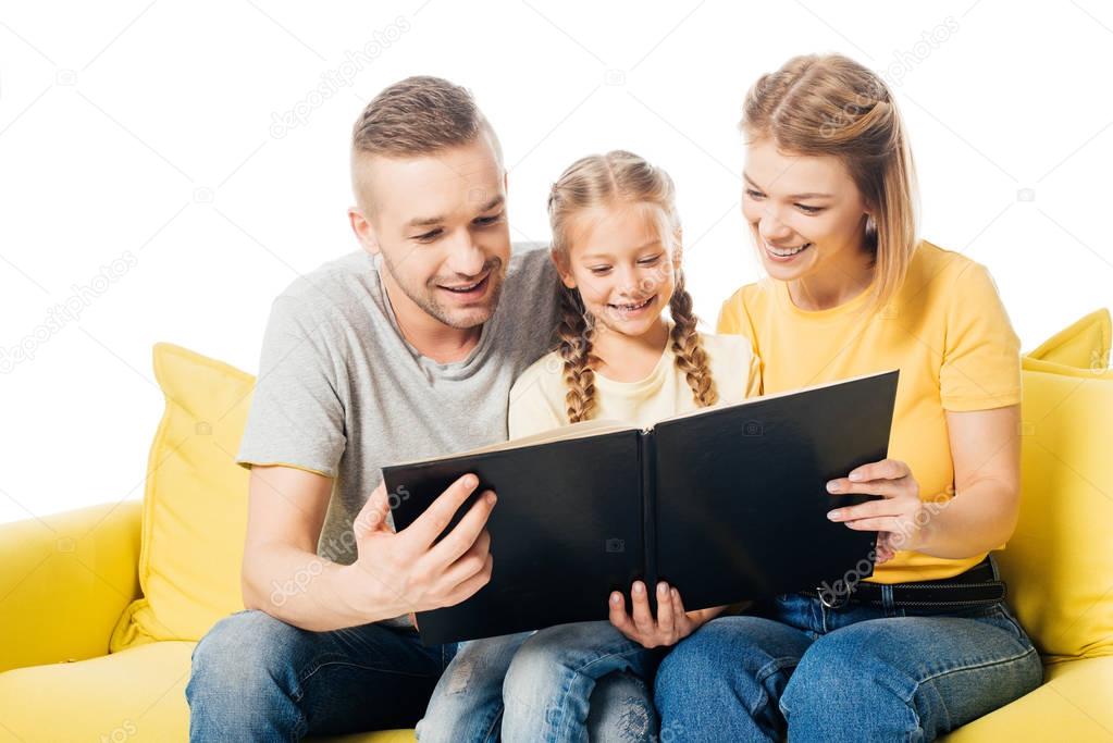 happy family looking at photos in photo album together while sitting on yellow sofa isolated on white