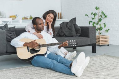 african american boyfriend sitting on floor and playing guitar for girlfriend at home clipart
