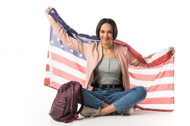Afrika kökenli Amerikalı öğrenci sırt çantası ile katta oturan ABD bayrağı tutarak gülümseyen, üzerinde beyaz izole 