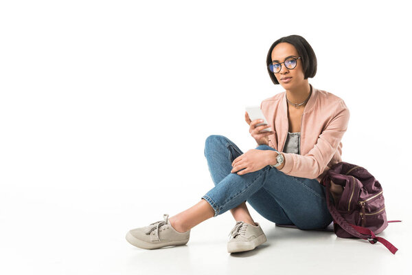 female african american student using smartphone while sitting on floor with backpack, isolated on white