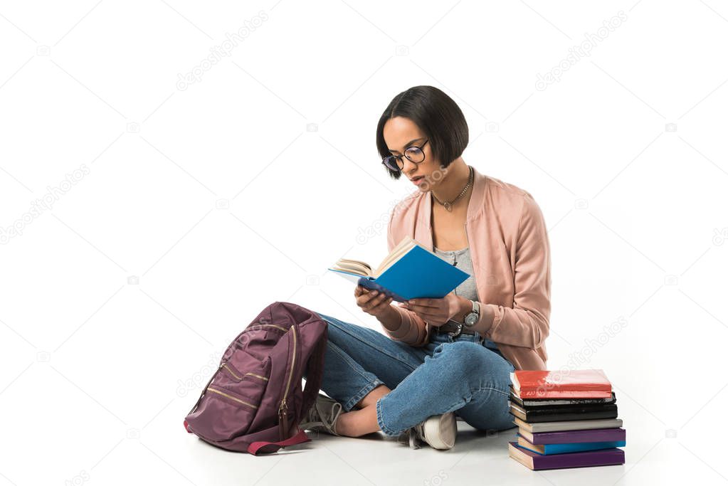 female african american student reading books while sitting on floor with backpack, isolated on white 