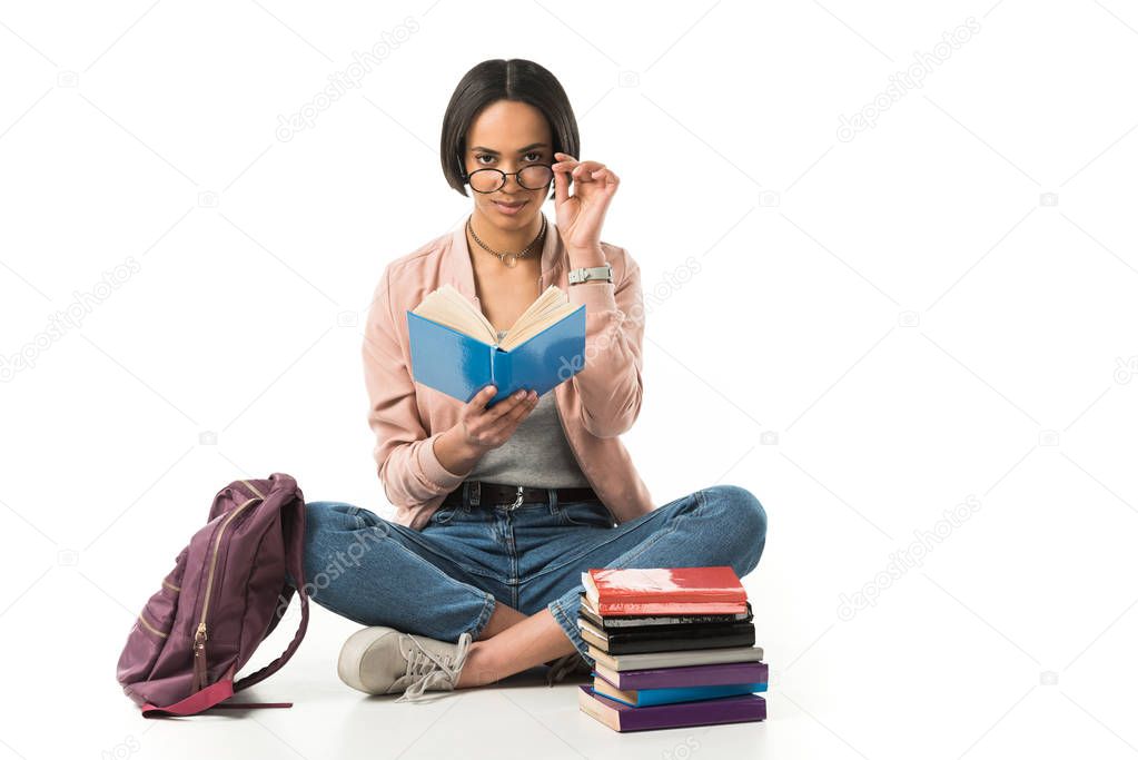 female african american student in glasses reading books while sitting on floor with backpack, isolated on white 