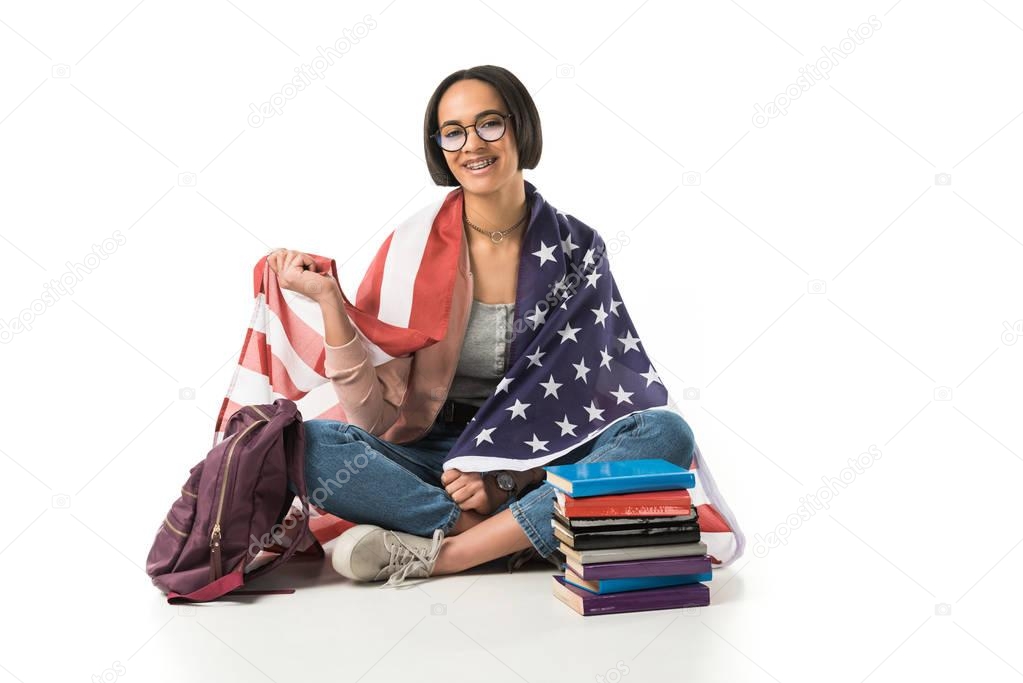 female african american student wrapped in usa flag sitting on floor with backpack and books, isolated on white 