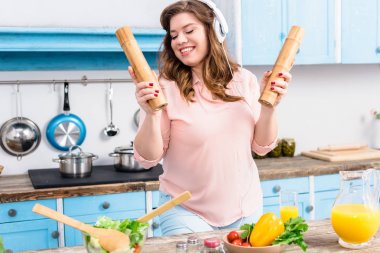 cheerful overweight woman in headphones with wooden salt and pepper grinders in hands in kitchen clipart