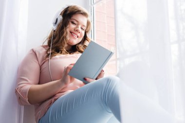 young woman in headphones reading book while sitting on windowsill at home clipart