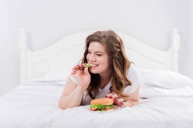 overweight woman in pajama eating burger on bed at home