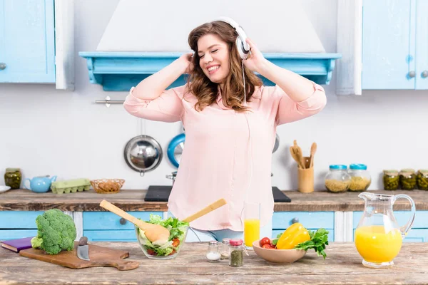 快乐超重妇女在家里厨房里用新鲜蔬菜在餐桌上听音乐 — 图库照片