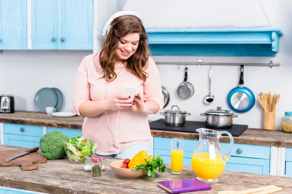 微笑超重妇女在耳机使用智能手机在餐桌上与新鲜蔬菜在家里厨房 — 图库照片