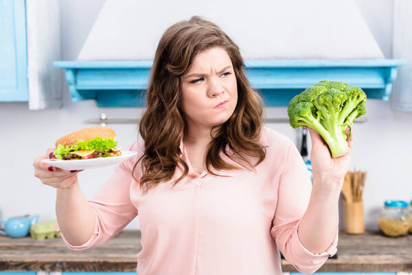портрет толстой женщины с бургером и свежей брокколи в руках на кухне дома, концепция здорового питания
