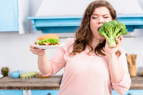 portrait of overweight woman with burger biting fresh broccoli in hands in kitchen at home, healthy eating concept