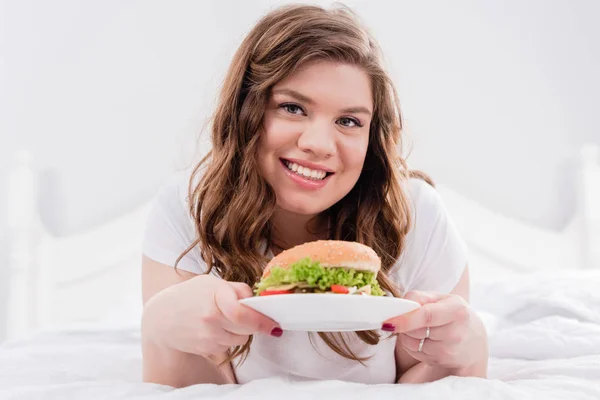 ハンバーガーを自宅のベッドの上でパジャマで太りすぎの笑顔の女性の肖像画 — ストック写真