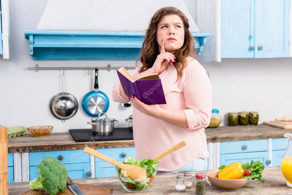 overweight pensive woman with cookery book standing in kitchen at home