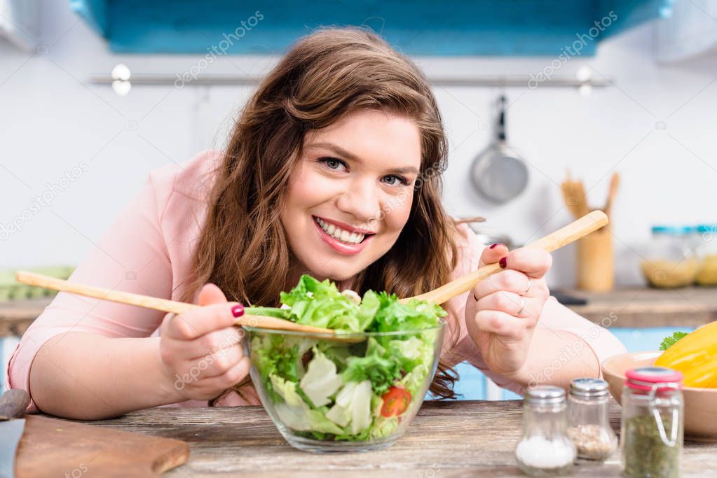 portrait of overweight smiling woman looking at camera while cooking fresh salad for dinner in kitchen at home