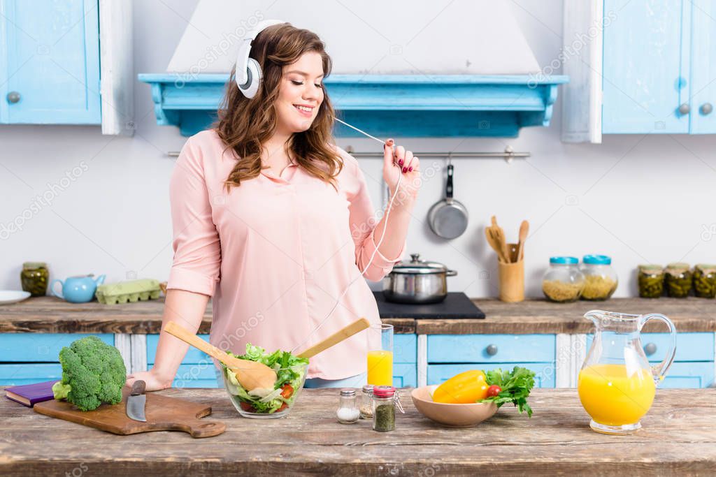 cheerful overweight woman listening music in headphones at table with fresh vegetables in kitchen at home