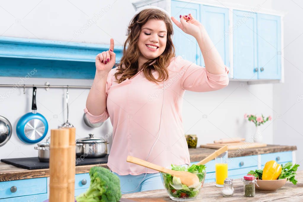 cheerful overweight woman listening music in headphones and dancing at table with fresh vegetables in kitchen at home