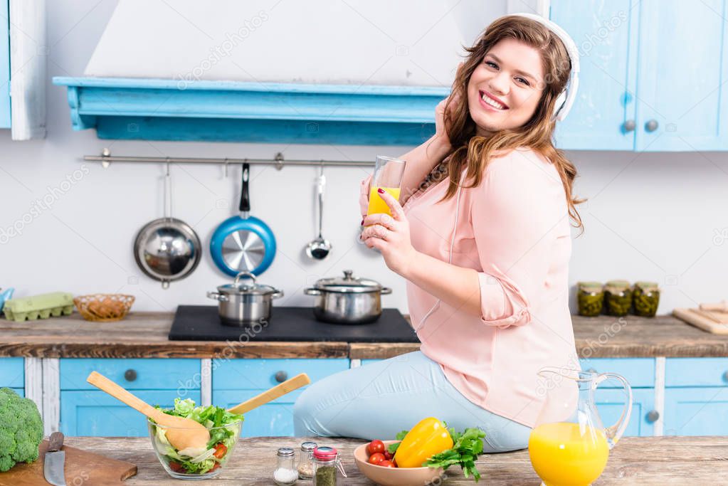 smiling overweight woman in headphones with glass of juice in hand in kitchen