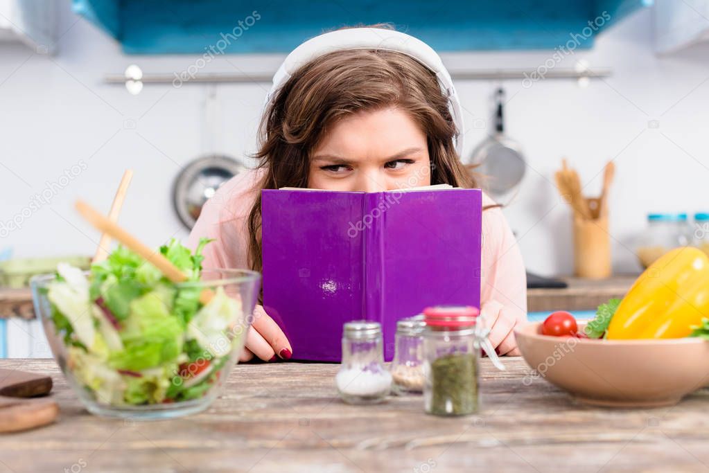 obscured view of overweight woman in headphones hiding behind cookery book at table with fresh salad in kitchen at home