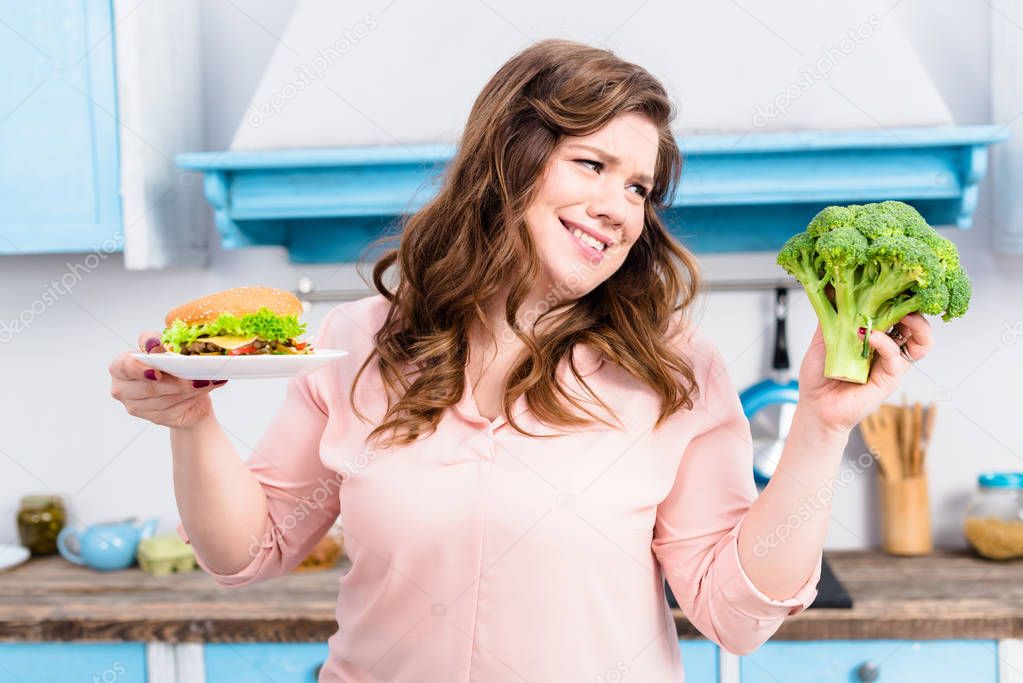 portrait of overweight woman with burger and fresh broccoli in hands in kitchen at home, healthy eating concept