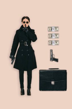 top view of female killer in sunglasses doing shushing gesture with briefcase, handguns and cash isolated on pink background clipart