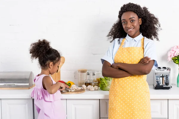 african american mom standing with crossed arms while daughter cutting vegetables on kitchen