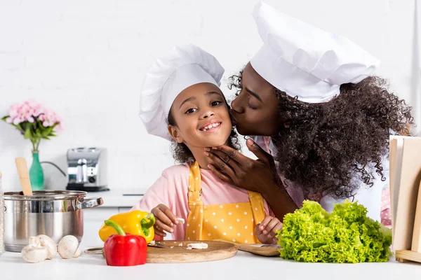 キッチンで料理をしながらシェフ帽子で彼女の娘をキス アフリカ系アメリカ人の母 — ストック写真