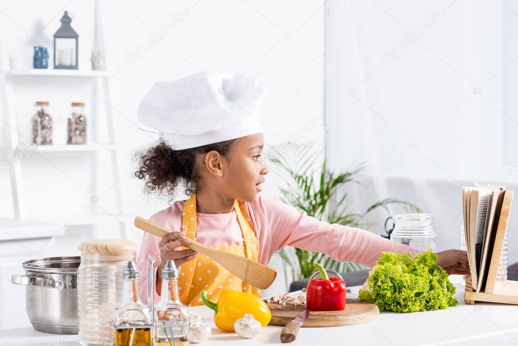 adorable african american child in apron and chef hat cooking on kitchen