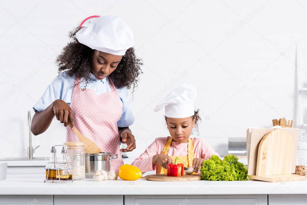 african american mother and daughter in chef hats cooking on kitchen