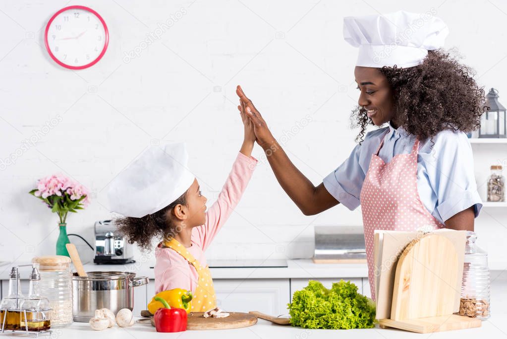 african american mother and daughter in chef hats cooking and giving high five on kitchen
