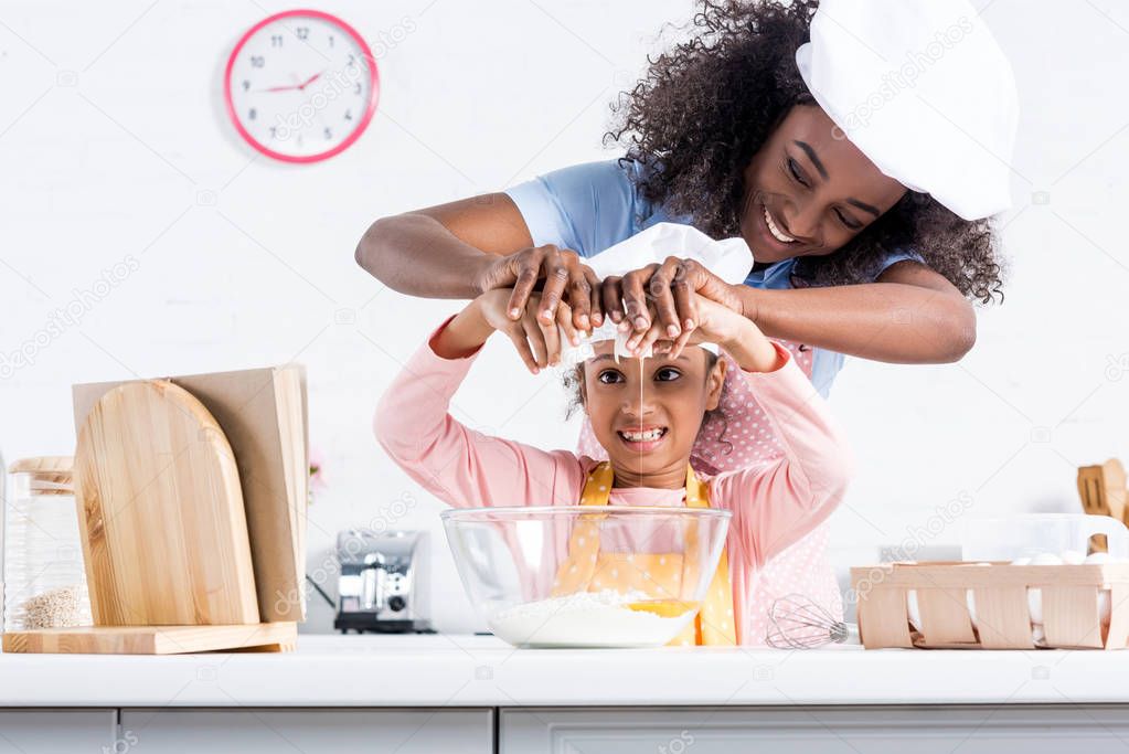 smiling african american mother and daughter in chef hats putting eggs into bowl together on kitchen