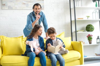 sessizlik için el hareketi ve kamera çocuklar evde kanepede kitap okuma sırasında bakarak mutlu baba