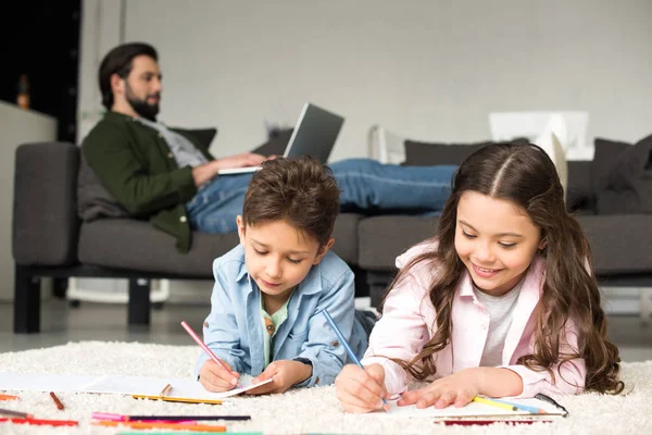 可爱的微笑的孩子躺在地毯上 用彩色铅笔画画 而父亲使用笔记本电脑在后面的沙发上 — 图库照片