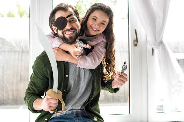 happy father and daughter smiling at camera while playing together and pretending to be pirates at home 