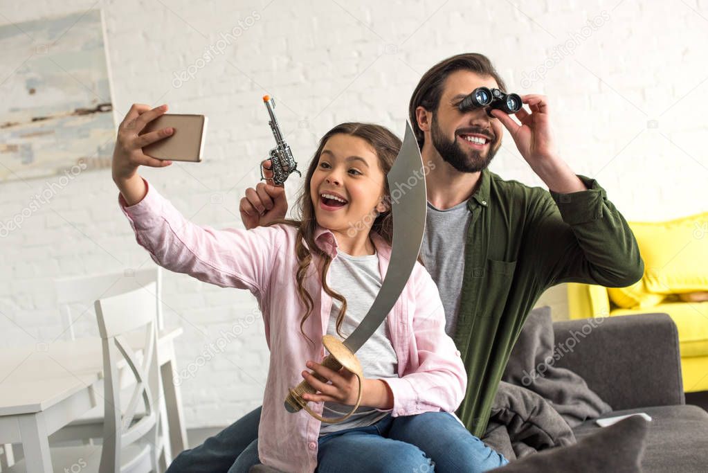 happy father and daughter pretending to be pirates and taking selfie with smartphone at home