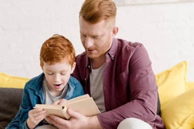 father looking at cute little son reading book at home clipart