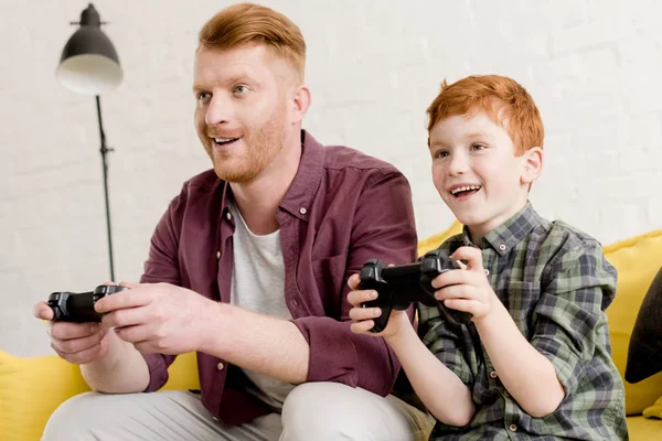 smiling father and son sitting on sofa and playing with joysticks at home