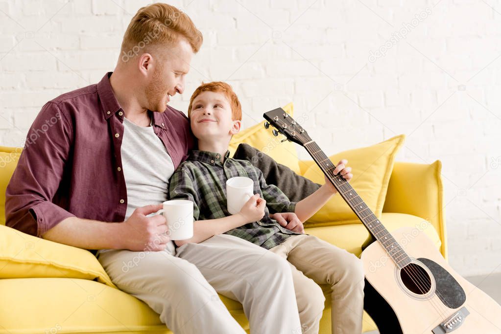happy father and son holding mugs and acoustic guitar while sitting on couch at home