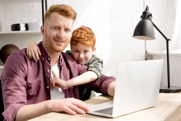 happy father and son smiling at camera while using laptop together at home