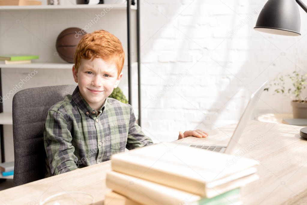 cute little redhead boy smiling at camera while sitting at table with books and laptop