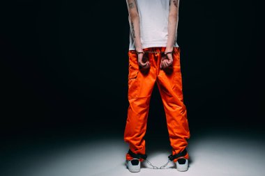 Cropped view of man wearing prison uniform with hands cuffed behind his back on dark background clipart