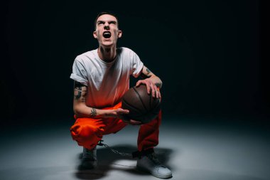 Screaming man in prison uniform and cuffs holding basketball ball on dark background clipart