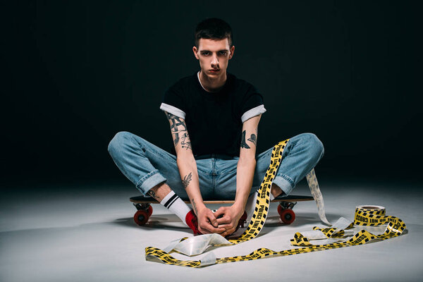 Hipster man sitting on longboard in police tape on dark background