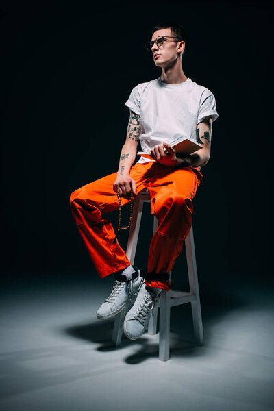 Man with tattoos wearing orange pants holding rosary and bible while sitting on stool on dark background